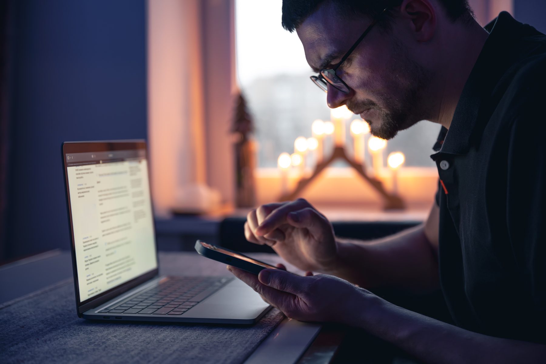 A man at a desk in the evening, laptop open, absorbed in his phone.